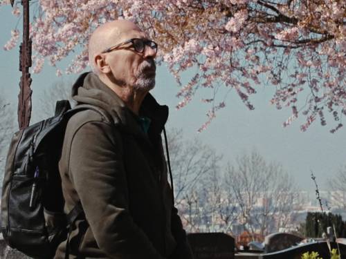 Man sieht einen Mann mit Halbglatze und dunkler Brille, welcher einen Rucksack trägt. Im Hintergrund blüht ein Kirschbaum. Anhand des unteres Bildausschnitts erkennt man, dass der Mann sich auf einem Friedhof befindet.